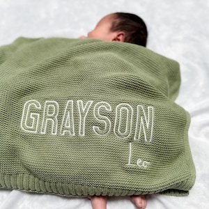 K&ouml;nnte beinhalten: Eine salbeigr&uuml;ne Babydecke aus Strick mit dem Namen "GRAYSON" in Wei&szlig; gestickt, darunter der Name "Leo". Die Decke ist &uuml;ber einem Baby drapiert.