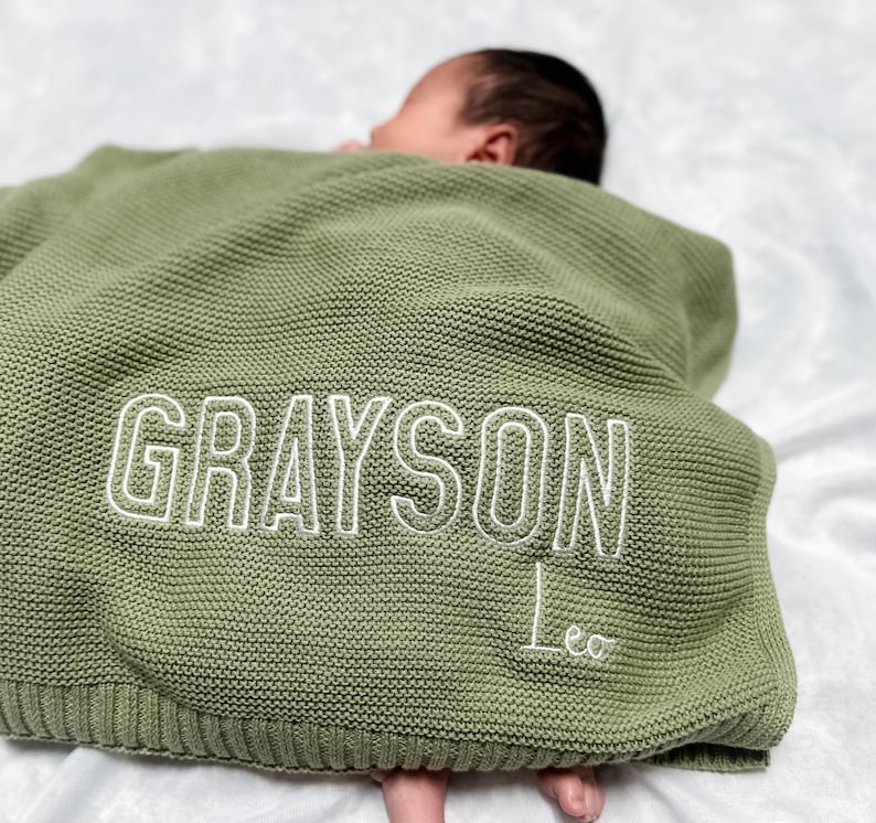 K&ouml;nnte beinhalten: Eine salbeigr&uuml;ne Babydecke aus Strick mit dem Namen "GRAYSON" in Wei&szlig; gestickt, darunter der Name "Leo". Die Decke ist &uuml;ber einem Baby drapiert.