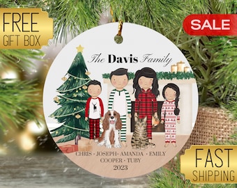 May include: A personalized Christmas ornament featuring a family of five in pajamas standing in front of a fireplace with a Christmas tree. The ornament is decorated with the text "The Davis Family", "Chris, Joseph, Amanda, Emily, Cooper, Tuby", and "2023".