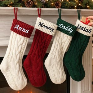 May include: Four cable-knit Christmas stockings in cream, red, and green hang from a mantelpiece. Each stocking has a contrasting cuff and is personalised with a name: Anna, Mommy, Daddy, and Isaiah. A hand places a small gift in one.