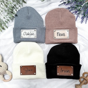 Peut inclure: Quatre bonnets en tricot avec des patchs en cuir personnalisés. Les bonnets sont en gris, rose, blanc et noir. Les patchs portent les noms Oaklan, Nova, Stone et Irvin.