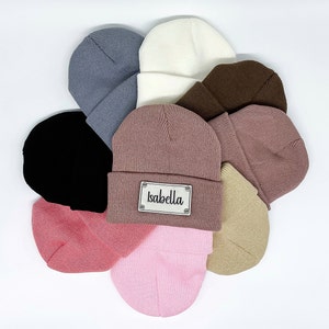 May include: A collection of ten knit beanies in various colors, including black, pink, brown, beige, and gray. One beanie has a white label with the name "Isabella" written in black.