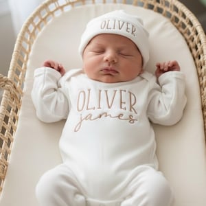 May include: A newborn baby wearing a white onesie and matching cap, both embroidered with the name "OLIVER" in a soft brown thread. The baby is lying in a woven basket, creating a serene and comforting scene.