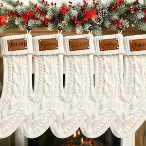 Peut inclure: Cinq chaussettes de Noël en tricot torsadé blanc cassé suspendues en rangée. Chaque chaussette comporte un écusson en cuir marron sur le revers supérieur, fixé par des rivets métalliques. Les noms McCoy, Madison, Beckham, Santiago et Chance sont gravés.