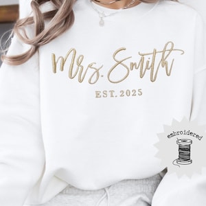 May include: A white sweatshirt with gold embroidered text that reads "Mrs. Smith" above "EST. 2025". The sweatshirt has a crew neck and long sleeves. A small embroidered label with a spool of thread is on the lower right.