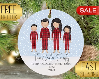 May include: A white round Christmas ornament with a blue background and a snowy design. The ornament features a family of five in red and black plaid pajamas. The text "The Carter Family" is written below the family, along with the names "Chris, Amanda, Rose, John, Lexi" and the year "2023".