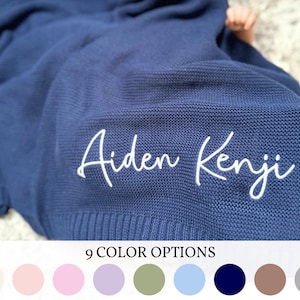 May include: A navy blue knitted baby blanket with the names "Aiden Kenji" embroidered in white script. The blanket is folded, with a baby's arm visible. The bottom of the image shows nine color options.