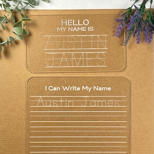 May include: Two clear acrylic name practice boards with the name "Austin James" written on them. The top board says "Hello My Name Is" and the bottom board says "I Can Write My Name".
