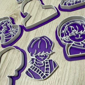 Pode incluir: Uma cole&ccedil;&atilde;o de cortadores de biscoitos de pl&aacute;stico roxo e branco em v&aacute;rias formas, incluindo um rosto e uma nuvem. Os cortadores est&atilde;o dispostos sobre uma superf&iacute;cie de madeira clara.