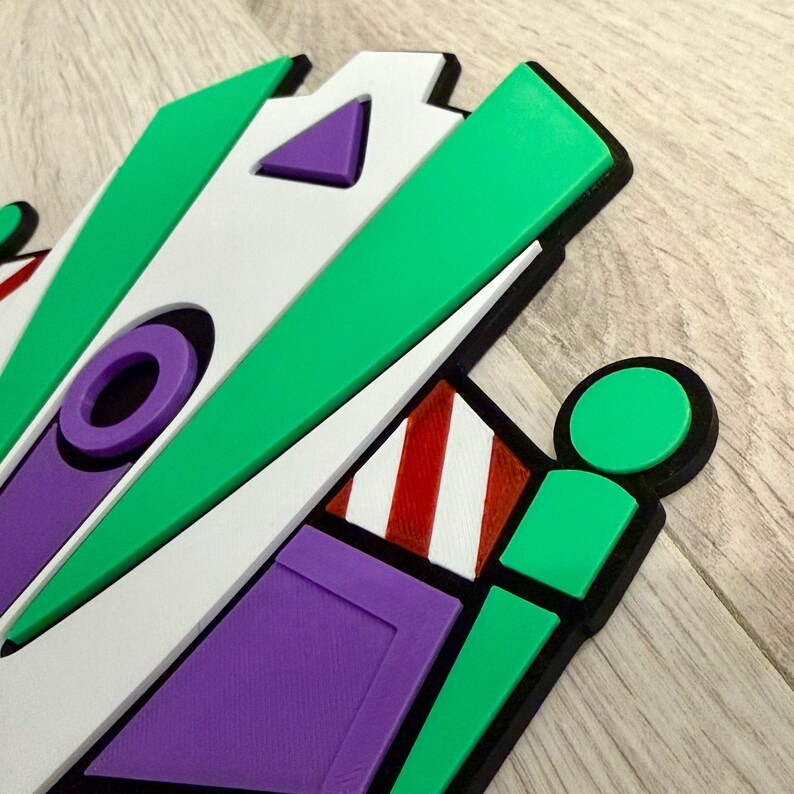 May include: A colorful, geometric design featuring green, white, purple, and red accents. The design has a black border and appears to be a stylized representation of a character or object. The design is on a light wood surface.