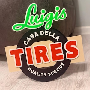Può includere: Un'insegna con la scritta "Luigis" in verde e bianco, sopra un'insegna che dice "Casa Della Tires Quality Service" in rosso, bianco e nero. L'insegna è su uno sfondo beige.