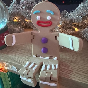 Pode incluir: Um enfeite de Natal de homem de gengibre castanho com sobrancelhas de glacê azul, boca vermelha e botões roxos. Os braços e as pernas são articulados. Detalhes de glacê branco contornam as bordas. Uma argola no topo para pendurar.