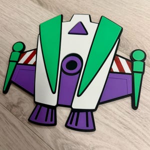 May include: A colorful, stylized depiction of a space shuttle, featuring green, white, purple, and black accents. The design includes wings, a central body, and thrusters, with red and white striped details. The overall shape is reminiscent of a cartoon character.