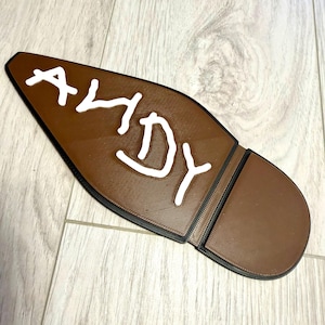 Puede incluir: Objeto marrón con forma de suela de zapato con el nombre "ANDY" escrito en letras blancas y estilizadas. El objeto tiene una superficie texturizada y un borde negro. El talón es de un tono marrón más oscuro.