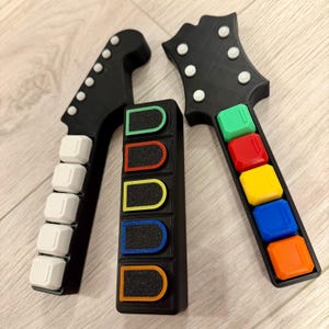 May include: Black guitar controllers with colourful buttons. The controllers have white fret buttons and colourful buttons in green, red, yellow, blue, and orange. The guitar heads have white circular inlays.
