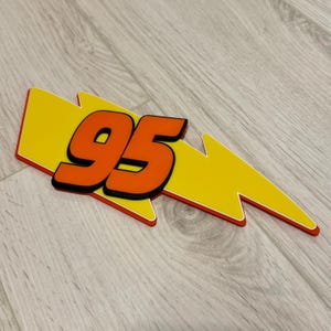 May include: A yellow lightning bolt shape with the number 95 in orange. The number has a black outline, and the lightning bolt has a red outline. The design is likely a decorative item or emblem.