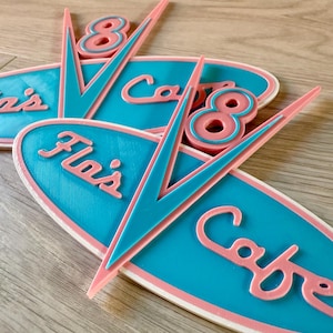 May include: A collection of retro cafe signs in teal, pink, and white. The signs display the text "Fla's Cafe" and the number "8". The design has a mid-century modern aesthetic, reminiscent of classic diners.