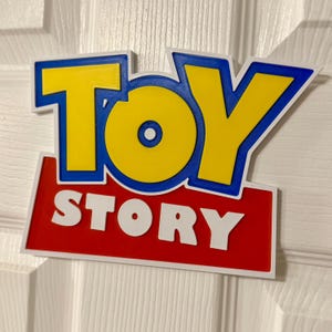 May include: A colorful sign featuring the words "TOY STORY" in bold, stylized lettering. The word "TOY" is in yellow with a blue outline, and "STORY" is in white on a red background. The sign is likely a decorative item or a piece of merchandise.