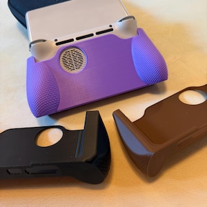 Puede incluir: Un agarre púrpura para consola de juegos portátil con agarres texturizados y una rejilla para el altavoz. También se ven dos fundas protectoras adicionales, una negra y otra marrón. La consola está parcialmente insertada en el agarre púrpura.