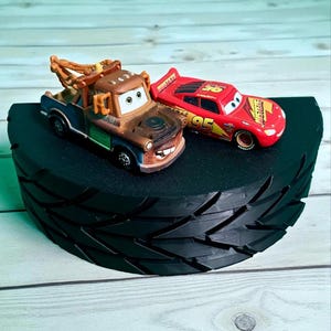 以下が含まれることがあります： 黒いタイヤ型のディスプレイスタンドには、茶色のレッカー車と赤いレーシングカーの2台のおもちゃの車が置かれています。レッカー車には茶色の牽引アームがあり、レーシングカーには95番の数字が書かれています。