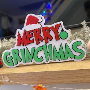 Puede incluir: Decoración navideña con las palabras "MERRY GRINCHMAS" en rojo y verde, con un gorro de Papá Noel y una bola verde. El diseño está sobre un fondo blanco con un borde blanco.