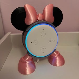 May include: A pink and black 3D printed stand for an Amazon Echo Dot smart speaker. The stand is designed to look like Mickey Mouse ears with a pink bow on top. The Echo Dot is visible in the stand with its blue light ring illuminated.