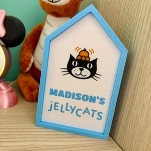 May include: A blue, house-shaped sign with the text "MADISON'S JELLYCATS" in blue. The sign features a black cat wearing a hat. A stuffed tiger and a Minnie Mouse figure are in the background.
