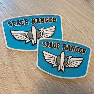 以下が含まれることがあります： 2つの青と白のスペースレンジャーのロゴプレート。各プレートには、翼、ロケット、惑星の様式化された画像の上に、黒い文字で「SPACE RANGER」という言葉が書かれています。プレートには白い縁があります。