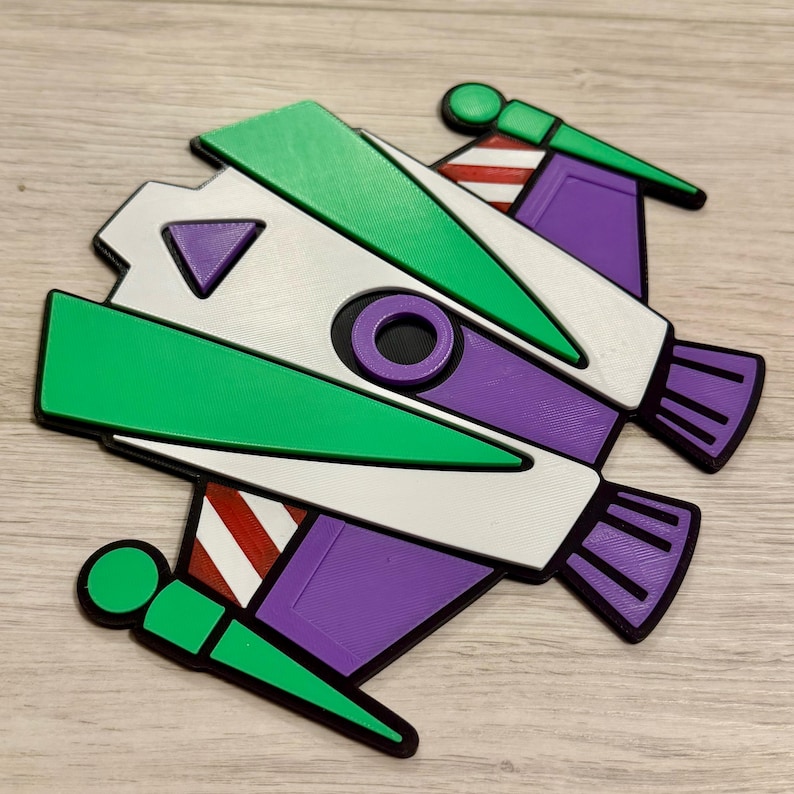 May include: A colorful, stylized 3D-printed spaceship toy in green, white, purple, and red. The toy features a circular design with wings and a rocket engine design. The edges are outlined in black.