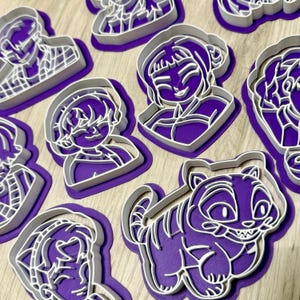 May include: A selection of purple and white plastic cookie cutters in diverse shapes, including smiling faces and a tiger. The cutters are displayed on a purple surface, highlighting their detailed designs and contrasting colours.