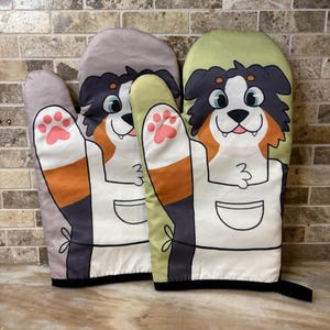 May include: Two oven mitts with a dog design. The mitts are light gray and light green. Each mitt features a cartoon dog with black, brown, and white fur, wearing a white apron. The dog's paws are raised, showing pink paw prints.