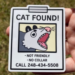 May include: A white sticker with a cartoon opossum illustration, framed by a black border. The text "CAT FOUND!" is at the top, with "- NOT FRIENDLY - NO COLLAR" below the illustration, and a phone number at the bottom.