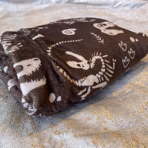 Op de afbeelding: Gevouwen bruine deken met een wit dinosaurus skeletpatroon. De deken is gemaakt van een zacht, pluche materiaal en heeft verschillende illustraties van dinosaurusbotten. De deken is netjes opgevouwen en toont het ontwerp.