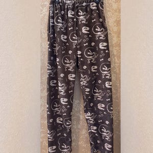 Op de afbeelding: Bruine pyjamabroek met een patroon van witte dinosaurus skeletten en botten. De broek heeft een elastische tailleband en een losse pasvorm. De stof ziet er zacht en comfortabel uit, perfect om in te loungen of te slapen.