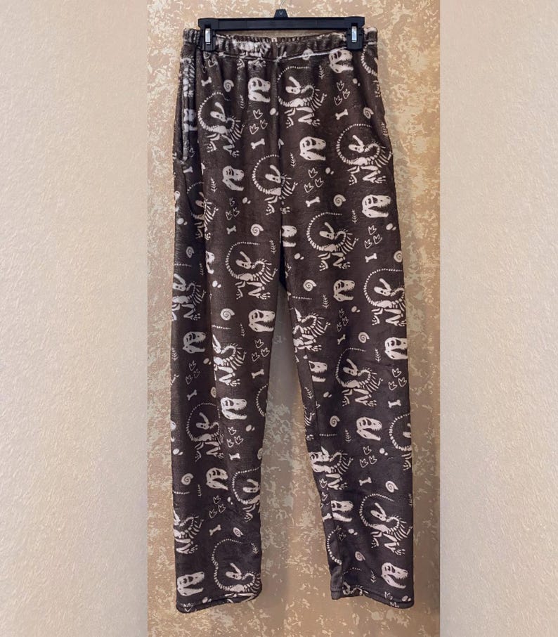 Op de afbeelding: Bruine pyjamabroek met een patroon van witte dinosaurus skeletten en botten. De broek heeft een elastische tailleband en een losse pasvorm. De stof ziet er zacht en comfortabel uit, perfect om in te loungen of te slapen.