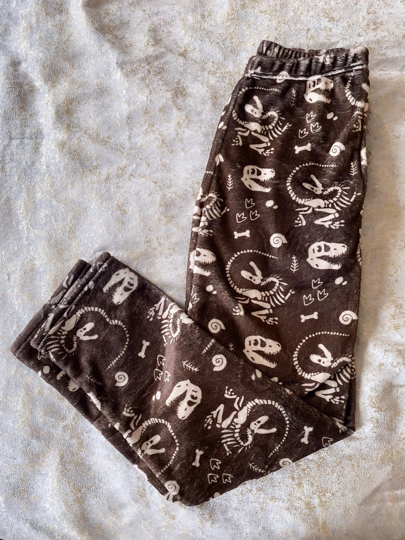 Op de afbeelding: Bruine pyjamabroek met een herhaald patroon van witte dinosaurus skeletten, botten en spiraalvormen. De broek heeft een elastische tailleband en is gemaakt van een zacht, pluche materiaal. Perfect om in te loungen of te slapen.