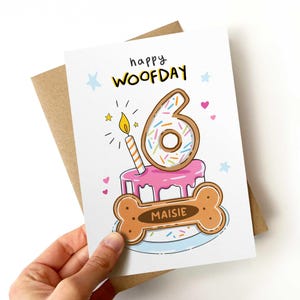 May include: A birthday card with a cartoon illustration. The card features a pink cake with a number 6 donut on top, a lit candle, and a dog bone with the name "Maisie". The words "happy WOOFDAY" are written at the top.