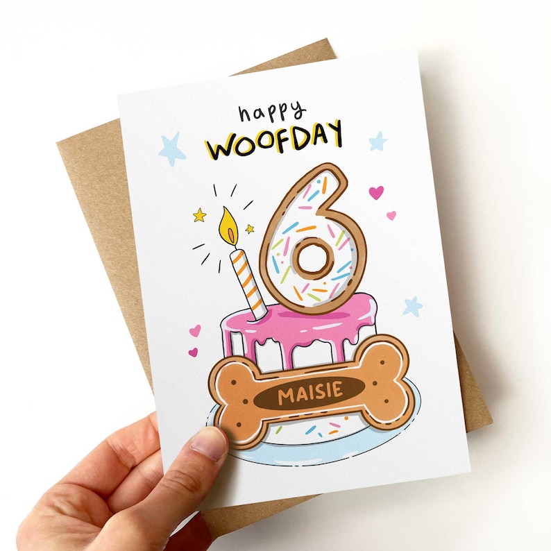 May include: A birthday card with a cartoon illustration. The card features a pink cake with a number 6 donut on top, a lit candle, and a dog bone with the name "Maisie". The words "happy WOOFDAY" are written at the top.