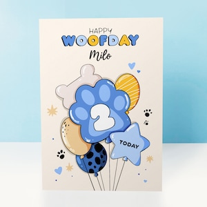 K&ouml;nnte beinhalten: Eine Geburtstagskarte mit einem Paw-Print-Ballon-Design. Die Karte ist cremefarben mit blauen, gelben und wei&szlig;en Paw-Print-Ballons. Die Ballons haben eine Nummer 2 und das Wort "TODAY" darauf. Die Karte sagt "Happy Woofday Milo".