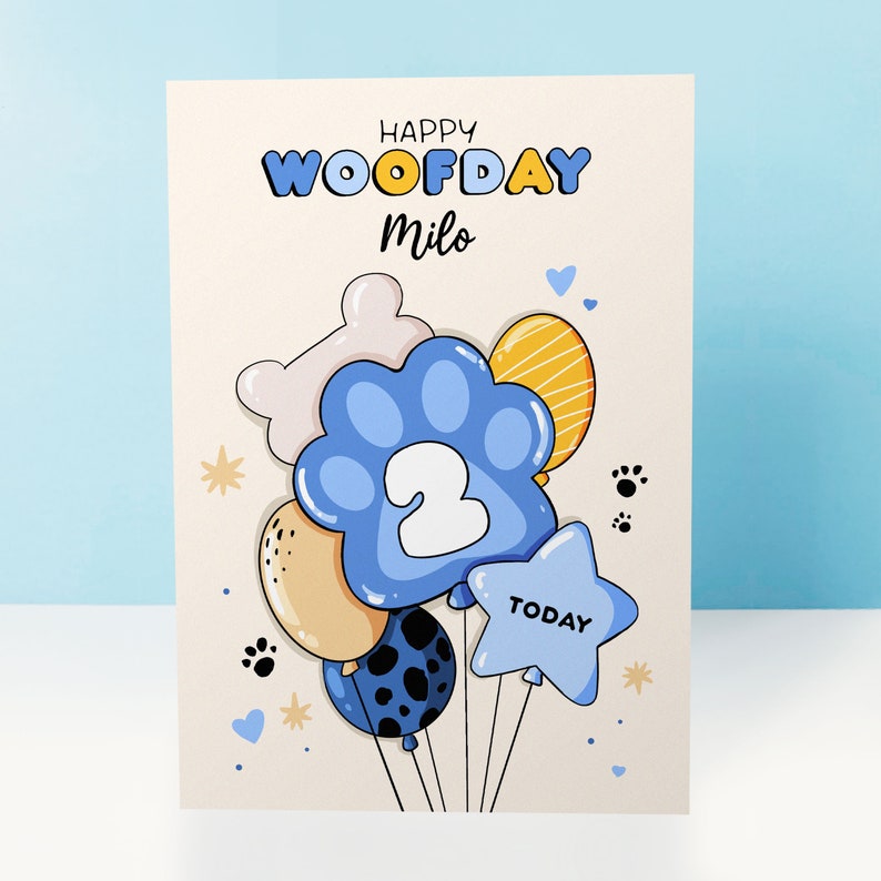 K&ouml;nnte beinhalten: Eine Geburtstagskarte mit einem Paw-Print-Ballon-Design. Die Karte ist cremefarben mit blauen, gelben und wei&szlig;en Paw-Print-Ballons. Die Ballons haben eine Nummer 2 und das Wort "TODAY" darauf. Die Karte sagt "Happy Woofday Milo".