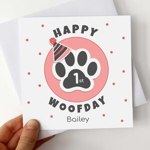 Puede incluir: Tarjeta de felicitaci&oacute;n cuadrada con fondo blanco y lunares rojos. Presenta un c&iacute;rculo rosa con una huella de pata negra y un gorro de fiesta. Las palabras "HAPPY WOOFDAY 1st" est&aacute;n impresas en la tarjeta, junto con el nombre "Bailey".