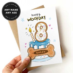 May include: A birthday card with a dog bone and cake design. The card says "happy WOOFDAY" and features a number 8 donut with sprinkles and a lit candle. The dog bone has the name "BAILEY" on it. The card is held in a hand.