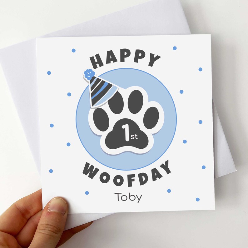 Puede incluir: Tarjeta cuadrada con un dise&ntilde;o azul y blanco. Presenta una huella de pata con un gorro de fiesta, las palabras "HAPPY WOOFDAY 1st" y el nombre "Toby". La tarjeta se sostiene frente a un sobre blanco.