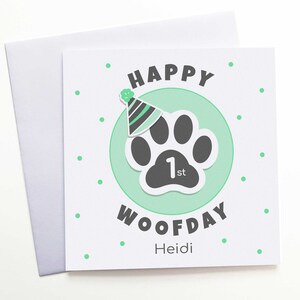 Puede incluir: Una tarjeta de felicitaci&oacute;n cuadrada blanca con un sobre a juego. La tarjeta presenta una huella de pata gris dentro de un c&iacute;rculo verde claro, un gorro de fiesta y las palabras "HAPPY WOOFDAY 1st Heidi" en negro y verde. Peque&ntilde;os puntos verdes est&aacute;n dispersos.