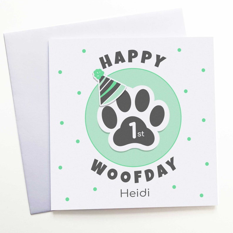 Puede incluir: Una tarjeta de felicitaci&oacute;n cuadrada blanca con un sobre a juego. La tarjeta presenta una huella de pata gris dentro de un c&iacute;rculo verde claro, un gorro de fiesta y las palabras "HAPPY WOOFDAY 1st Heidi" en negro y verde. Peque&ntilde;os puntos verdes est&aacute;n dispersos.