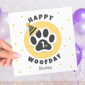 Puede incluir: Una tarjeta cuadrada blanca con el texto "HAPPY WOOFDAY" y un dise&ntilde;o de huella de pata. La huella de pata es negra con un fondo de c&iacute;rculo amarillo y un gorro de fiesta. La tarjeta tambi&eacute;n incluye el texto "1st" y el nombre "Bailey".