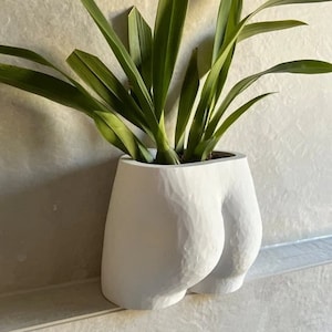 May include: A white ceramic planter, sculpted to resemble a human form, holding a green plant with long leaves. The planter has a textured surface and is set against a light-coloured wall.