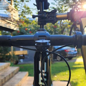 Puede incluir: Primer plano de un manillar de bicicleta de carretera negro con manetas de freno integradas. El manillar está envuelto en cinta negra. El cuadro de la bicicleta tiene el nombre de la marca "BMC" en letras blancas. El fondo es un entorno residencial soleado.