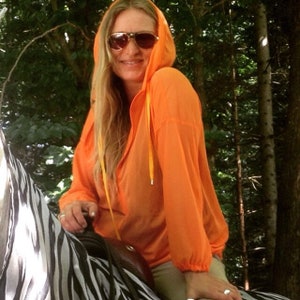 May include: A woman wearing a bright orange hooded jacket with a yellow drawstring. She is wearing sunglasses and has long blonde hair. She is sitting on a zebra-patterned saddle.