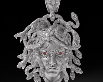 May include: A silver pendant featuring a detailed sculpture of Medusa, the Greek mythological figure with snakes for hair. Her face is sculpted with a fierce expression, and her eyes are red.